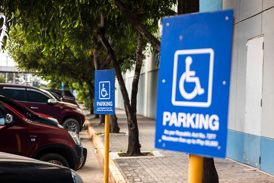 Two blue parking signs featuring the white wheelchair symbol indicating designated accessible parking spaces are positioned along a sidewalk next to a building with a light-colored wall. The signs are mounted on yellow poles, with the larger sign in the foreground and the smaller one further back. Behind the signs, there are several parked cars, including a dark-colored SUV, on a paved parking area adjacent to a tree with green leaves. The scene appears to be outdoors during daytime, with natural lighting illuminating the area. The setting suggests a public parking zone that might be relevant for home relocation or furniture transport activities, and the image supports understanding of parking regulations that could impact moving logistics, with signage visible from the pavement near an urban property. Man with Van Longlands may use such imagery to illustrate parking considerations during moves.