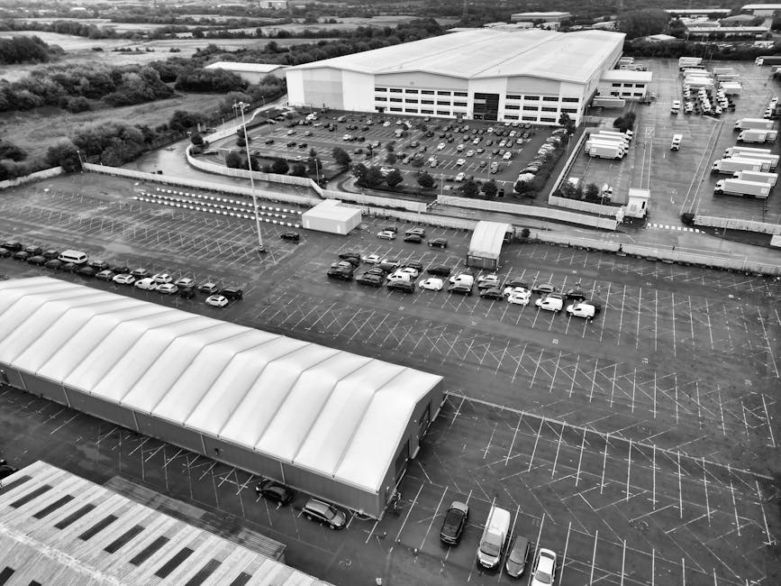 An aerial view of a large commercial parking lot adjacent to a warehouse or industrial building, with numerous parking spaces marked out with white lines, mostly unoccupied with a few cars parked in designated areas. Several trucks and trailers are parked along the side of the building, some near loading bays. The parking area includes a covered structure with a curved roof, likely used for vehicle storage or loading purposes, and a smaller white trailer or container positioned near the center of the lot. The environment appears to be in daytime with overcast lighting. The surrounding area features additional storage facilities or industrial units, with patches of greenery in the distance. The image relates to house removals and furniture transport, illustrating logistics and vehicle utilization relevant to packing, loading, and moving services provided by Man with Van Longlands, especially in contexts of home relocation or large-scale transfer.
