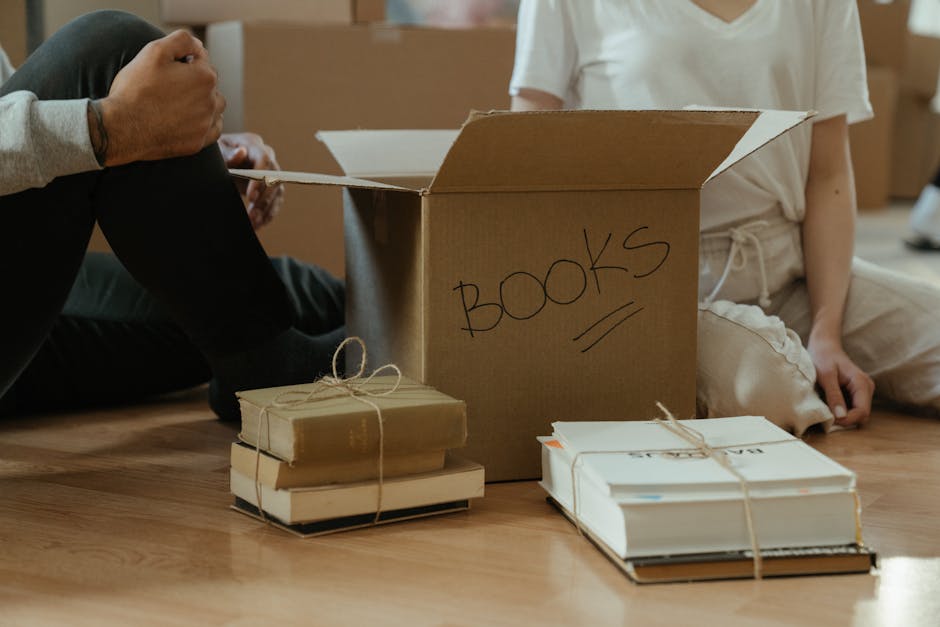 Two individuals are seated on a wooden floor, engaged in packing or unpacking household belongings as part of a home relocation process. In front of them, there is a large open cardboard box labeled '