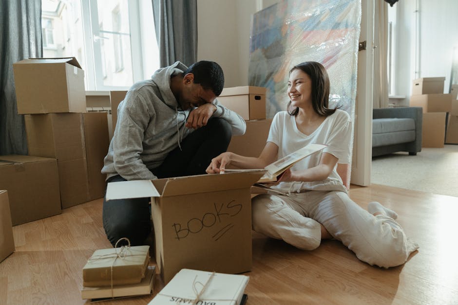 Two individuals are seated on a wooden floor, engaged in packing or unpacking household belongings as part of a home relocation process. In front of them, there is a large open cardboard box labeled '