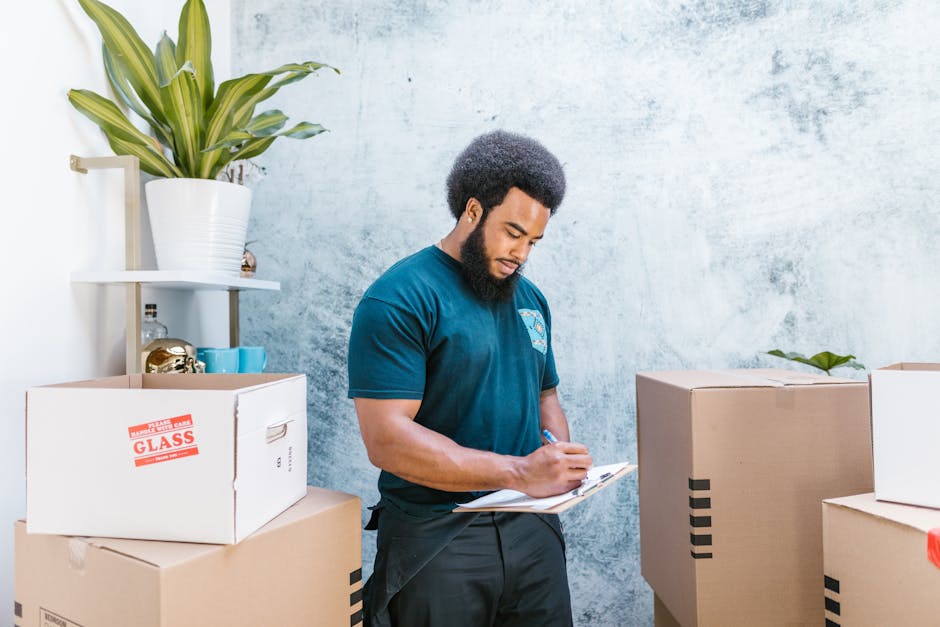 A man with dark curly hair and a beard, wearing a navy blue t-shirt, is standing indoors surrounded by moving boxes, some labeled with red stickers for fragile items like glass. He is holding a clipboard and a pen, appearing to review or check off items during a home relocation process. The boxes are made of cardboard, with some wrapped in plastic for protection, and are positioned on the floor and a nearby surface. Behind him, there is a white wall with a small shelf holding a large green potted plant, some decorative objects, and other small containers. The scene takes place in a well-lit interior space, possibly part of the packing and moving preparations carried out by a professional removals service such as Man with Van Longlands. The environment and objects suggest a professional and organized approach to furniture transport and home relocation, with an emphasis on careful handling of belongings during packing and loading.
