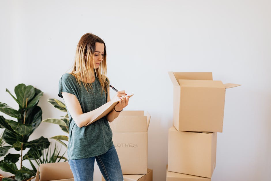 A young woman with long blonde hair, wearing a casual grey t-shirt and blue jeans, is standing indoors amidst several cardboard moving boxes. She is holding a clipboard and a pen, appearing to take notes or check off a list related to her home relocation process. The boxes, some open and others closed, are stacked on the floor, with one large box partially open revealing packing paper inside. To the left, a large green houseplant adds a touch of decor to the plain white wall background. The lighting is natural, illuminating the scene clearly. This setting reflects the packing and moving phase of a house removal, with a focus on the organization and documentation needed before a furniture transport or relocation, similar to procedures followed by services like Man with Van Longlands when managing damage claims after a move.