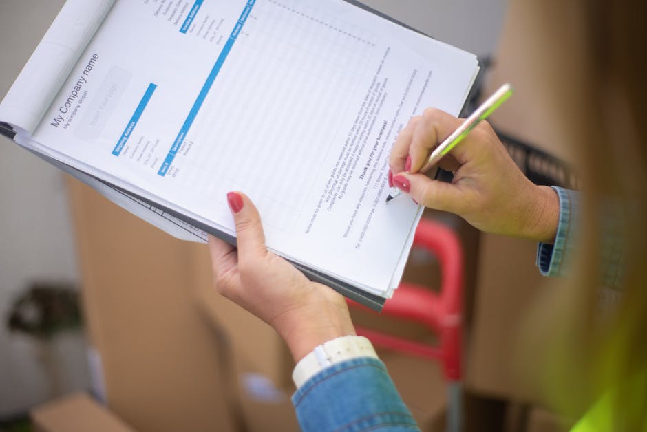 A person wearing a denim jacket and a white wristwatch is holding a clipboard with several pages of documents, including a form titled 'My Company Name.' They are using a pink and green pen to fill out or review the paperwork. The background shows part of a room with a cardboard box and a blurry red trolley, suggesting a home or office moving environment. The scene indicates involvement in logistics or paperwork related to house removals or relocation services, with the focus on the detailed documentation process. This image aligns with the packing and moving processes of [COMPANY_NAME], supporting the topic of whether permits are needed for Longlands removals according to council rules.