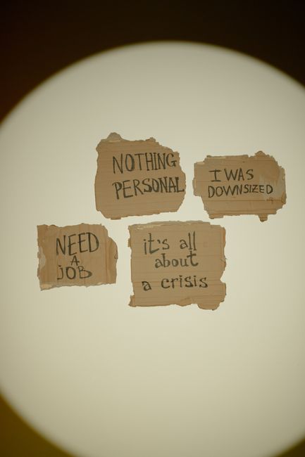 A close-up view through a circular viewing window reveals four torn pieces of brown paper taped onto a plain, off-white wall, each displaying handwritten messages in black ink. The messages read, 'NOTHING PERSONAL,' 'I WAS DOWNSIZED,' 'NEED A JOB,' and 'it's all about a crisis.' The torn edges of the paper suggest a casual or urgent display, with the focus on personal struggles related to job loss or economic hardship. No furniture or moving equipment is visible in the image, and the lighting is soft and diffused, creating a neutral environment. This visual could relate to the emotional side of home relocations or the challenges faced during moving and packing, which Man with Van Longlands handles during residential removals and moving services.