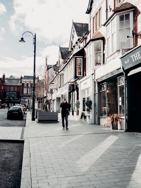 A man dressed in dark clothing walking along a wide, paved sidewalk outside a row of narrow, multi-storey retail shops with large glass windows and white facades on Longlands High Street. The shops are adorned with potted plants and decorative elements, and some have signage indicating businesses such as clothing stores or cafes. A black van is parked on the street to the left, and a black lamppost with a curved arm holding a streetlight is visible. The sky is partly cloudy with some blue showing, and the scene is well-lit, suggesting daytime. The image captures a typical high street scene that could relate to home or furniture relocation, with the presence of a moving company like Man with Van Longlands involved in packing or transporting belongings during a house move or local delivery process.