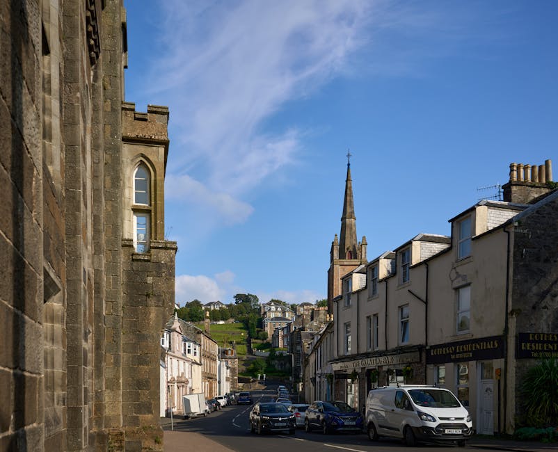 Photograph of a narrow urban street in Longlands with classic stone and brick buildings on both sides, some featuring small shops and residential entrances, under a clear blue sky with scattered clouds. In the distance, there is a tall church steeple rising above the rooftops, and a hillside with greenery and additional houses visible beyond. Parked cars and a white van are situated along the curb, indicating regular traffic and loading activity. The scene captures a typical small town street scene that may require careful navigation during a home relocation, with potential for narrow-access moving challenges that Man with Van Longlands can assist with during furniture transport and packing and moving operations.