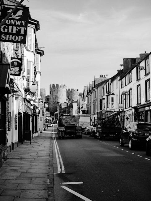 A man dressed in dark clothing walking along a wide, paved sidewalk outside a row of narrow, multi-storey retail shops with large glass windows and white facades on Longlands High Street. The shops are adorned with potted plants and decorative elements, and some have signage indicating businesses such as clothing stores or cafes. A black van is parked on the street to the left, and a black lamppost with a curved arm holding a streetlight is visible. The sky is partly cloudy with some blue showing, and the scene is well-lit, suggesting daytime. The image captures a typical high street scene that could relate to home or furniture relocation, with the presence of a moving company like Man with Van Longlands involved in packing or transporting belongings during a house move or local delivery process.