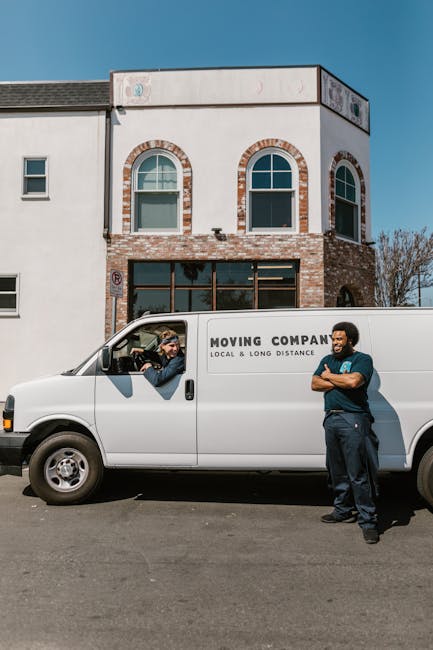 A white moving van parked on the street in front of a modern, multi-story residential building with a white and brick facade, large windows, and an arched design. Inside the van, partially visible through the open driver’s side window, a person wearing a beanie and glasses is handing out a clipboard or document to a man standing outside. The man, with arms crossed and smiling, is dressed in a dark blue uniform, possibly a mover from Man with Van Longlands, part of a home relocation or furniture transport service. The scene is set during daytime with clear skies, and the van is positioned in an area designated for loading or unloading. Visible around the scene are the building's exterior walls, a small tree, and pavement, illustrating a typical urban or suburban environment suited to packing and moving activities associated with house removals near Longlands Station.