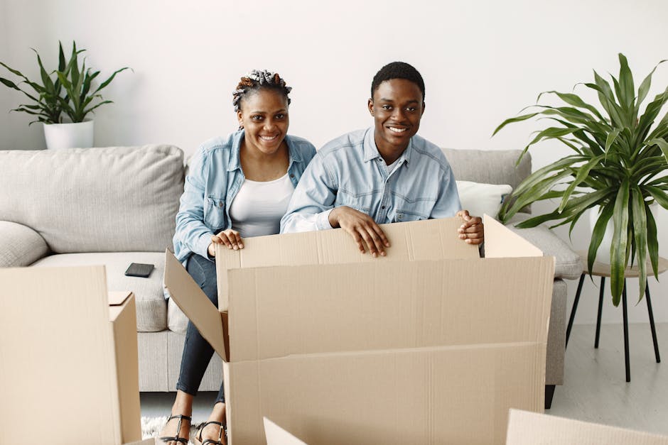A young couple with dark skin, smiling, preparing for a house move inside a well-lit living room. The woman, wearing a light blue denim jacket and white top, is sitting on a beige sofa with a remote control beside her. The man, also in a light denim shirt, is kneeling next to a large, open cardboard box on the floor, appearing ready to pack or unload belongings. Behind them, there are two potted green plants on either side of the sofa, contributing to a bright and welcoming atmosphere. The floor is light-colored wood, and the background features a plain white wall. The scene captures the initial stages of home relocation, involving packing items into boxes in a domestic setting. Man with Van Longlands frequently provides furniture transport and packing services to assist with house removals, emphasizing careful handling and organized moving processes.