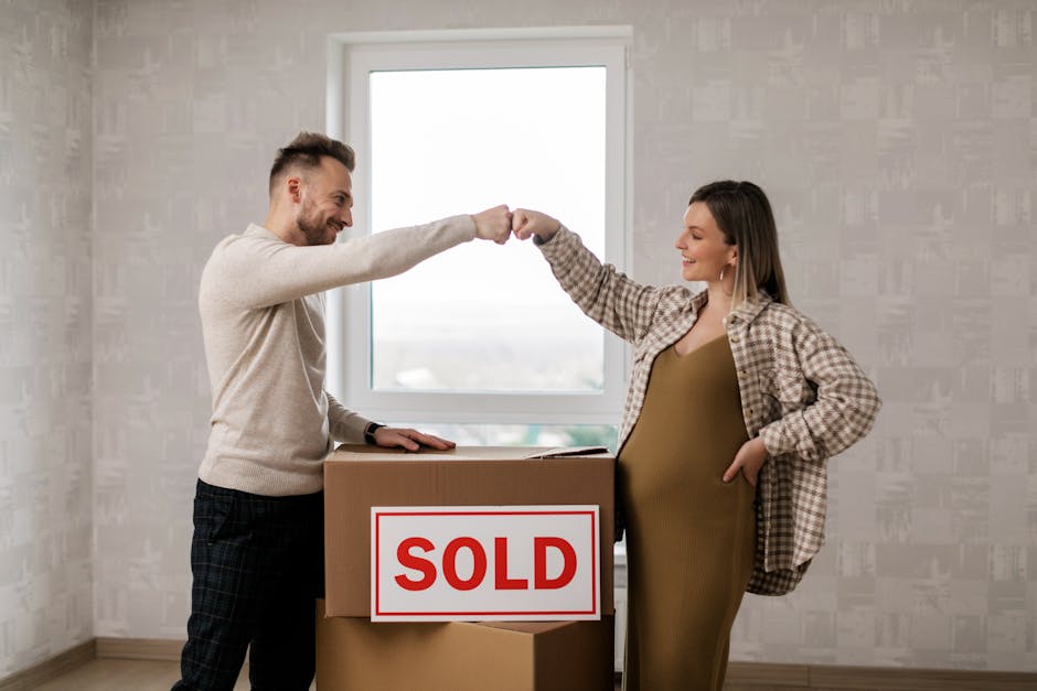 A man and a woman standing inside a new house during a home relocation process, engaging in a fist bump as a sign of successful sale and moving. The man, with short hair and a beard, is dressed in a light beige sweater and dark checkered trousers, resting one hand on a cardboard box with a prominent red and white 'SOLD' sign affixed to it. The woman, with shoulder-length hair, is wearing a plaid shirt layered over a mustard-colored dress, and has one hand in her pocket while the other is raised for a fist bump. Behind them, a wooden flooring and a plain, light-colored wall are visible, with a large window allowing natural daylight to illuminate the scene. The box and the 'SOLD' sign indicate they are in the process of packing or preparing for a move, symbolising a successful property sale and upcoming furniture transport for an estate move, which aligns with the services offered by [COMPANY_NAME], Man with Van Longlands, in house removals and relocation assistance.