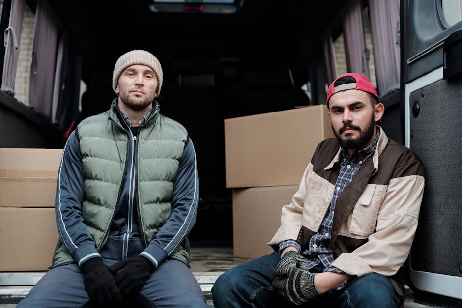 Two men sit on the floor inside the open rear of a van used for home relocation, with large cardboard boxes and packing materials such as plastic wrap and blankets stacked behind them. One man, on the left, wears a beige beanie, a green quilted vest over a dark hoodie, and black gloves, while the other, on the right, has a red cap, a beige and brown jacket, a checkered shirt, and gloves. Both men have serious expressions as they rest during the furniture transport process, which is part of a professional removal service by Man with Van Longlands. The environment is lit with natural daylight, revealing the interior of the van, which is set up for packing and loading household items, emphasizing the logistical aspect of house removals, packing, and furniture transport as seen in the service category.
