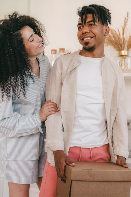 This image depicts a young man and woman indoors, likely within a residential property, involved in a home relocation process. The man is holding a medium-sized cardboard box with both hands, positioned at waist level, preparing for or involved in packing or moving furniture. He is dressed in a casual white t-shirt and light-colored jacket, with coral-colored trousers, and is smiling while looking away from the camera. The woman, wearing a striped shirt dress, stands close behind him, touching his shoulder and smiling at him, indicating a collaborative and positive atmosphere. The background includes a white wall with decorative items, such as a vase with dried pampas grass and picture frames, suggesting a domestic setting. The lighting is bright and natural, emphasizing the indoor environment. This scene captures the preparation phase of house removals, with a focus on packing and furniture transport, and is relevant to the services offered by Man with Van Longlands for short-notice student moves or other household relocations.