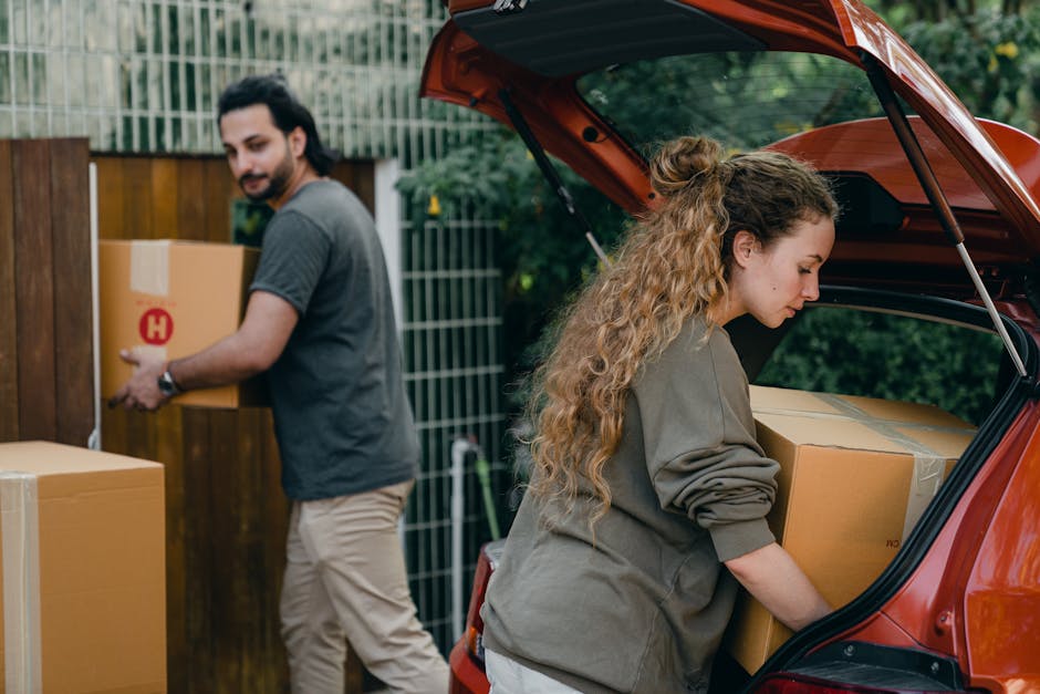 A woman with long, curly hair is placing cardboard boxes into the trunk of a red hatchback vehicle during a home relocation. She is wearing a grey sweatshirt and standing at the open rear of the car, carefully loading the boxes for transport. In the background, a man with dark hair and a beard, dressed in a grey t-shirt and beige trousers, is carrying a cardboard box towards a wooden fence and garden area. The setting appears to be an outdoor residential space with a green fence and lush foliage. The scene involves furniture transport and packing activities as part of a house removal process, with visible cardboard boxes and packaging materials, supported by the services of Man with Van Longlands as indicated by the webpage context.
