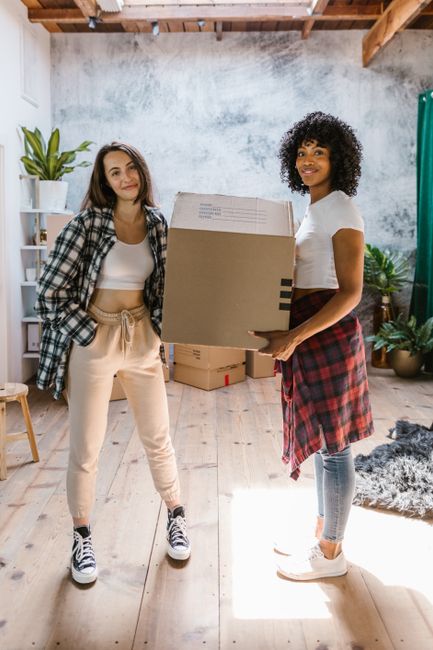 This image depicts a young man and woman indoors, likely within a residential property, involved in a home relocation process. The man is holding a medium-sized cardboard box with both hands, positioned at waist level, preparing for or involved in packing or moving furniture. He is dressed in a casual white t-shirt and light-colored jacket, with coral-colored trousers, and is smiling while looking away from the camera. The woman, wearing a striped shirt dress, stands close behind him, touching his shoulder and smiling at him, indicating a collaborative and positive atmosphere. The background includes a white wall with decorative items, such as a vase with dried pampas grass and picture frames, suggesting a domestic setting. The lighting is bright and natural, emphasizing the indoor environment. This scene captures the preparation phase of house removals, with a focus on packing and furniture transport, and is relevant to the services offered by Man with Van Longlands for short-notice student moves or other household relocations.