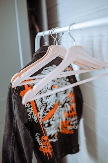 Close-up view of a white metal clothing rail mounted inside a property, holding several garments on white plastic hangers, including a black T-shirt with a large graphic print featuring white and orange elements. The clothing rail is positioned near a light-colored wooden wall, with part of a white wall or paneling visible in the background. The garments are arranged neatly, representing items typically prepared for packing or transportation during a home relocation. The scene suggests an organized packing process as part of furniture or house removal services offered by Man with Van Longlands, aligned with the process of loading items for a move in Longlands.