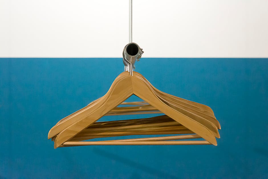 A set of five wooden clothes hangers are hanging on a metal hook attached to a wall with a blue and white horizontal division. The hangers are empty and uniformly arranged, with their curved sides facing outward. They are positioned inside a property, likely an interior closet or wardrobe area, with the hangers suspended in mid-air. The lighting is even, revealing the natural wood grain of each hanger. This setup suggests the process of packing or preparing clothing for a home relocation, which is part of the furniture transport and packing services offered by Man with Van Longlands. The image emphasizes the careful handling of garments and the organization involved in house removals, highlighting the logistics of loading and transporting belongings during a move.